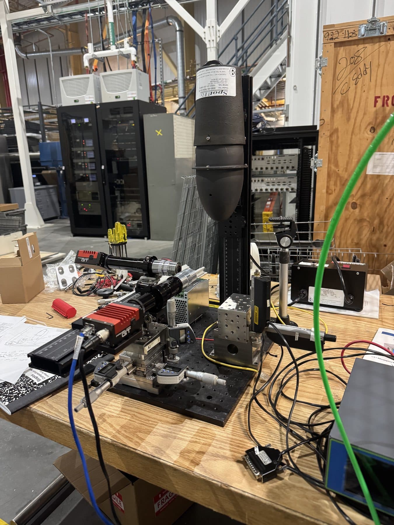 My bench at Brookhaven: the ellipsoidal reflector furnace mounted on a vertical rail above an optical breadboard, with two machine-vision cameras mounted on microscope lens tubes pointing at the sample position from different angles, a stack of micrometer translation stages, and instrument racks behind.